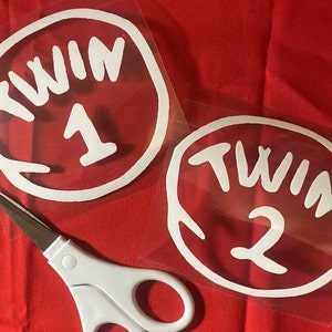 May include: Two white vinyl decals with the words "TWIN 1" and "TWIN 2" inside a white oval. The decals are on a clear backing, resting on a red surface. A pair of scissors with white handles is in the foreground.