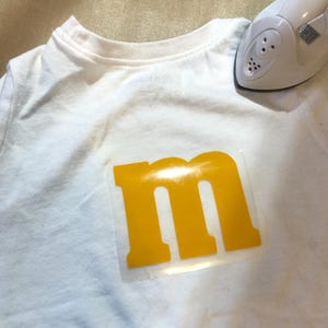 May include: A white t-shirt with a yellow letter "M" heat transfer vinyl design. A small white iron is positioned near the shirt's collar, suggesting the design is being applied. The shirt is on a light-colored surface.