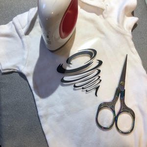 May include: A white onesie with a black vinyl design of a tornado on the front. A heat press and scissors are also in the image.
