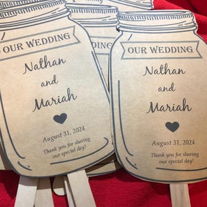 May include: Brown paper wedding fans shaped like mason jars with the text "Our Wedding" and the names "Nathan and Mariah" and the date "August 31, 2024". The fans have a small heart and the text "Thank you for sharing our special day!"