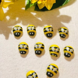 May include: Ten wooden bee-shaped ornaments painted yellow with black stripes and white wings. The bees have black eyes and are arranged in a circle on a white surface.