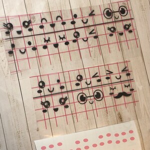 May include: A sheet of clear vinyl with black adhesive cutouts of various facial expressions, including eyes, mouths, noses, and eyebrows. The sheet is divided into a grid pattern with red lines.