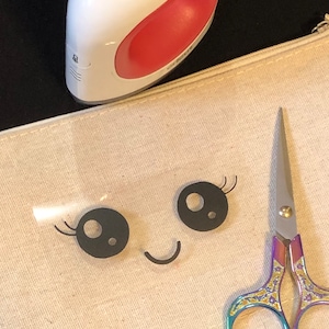 May include: A white heat press with a red handle is on top of a beige fabric pouch with a black and white cartoon face design. A pair of rainbow-colored scissors with gold accents is on the right side of the pouch.