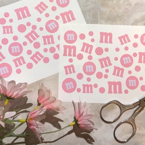 May include: Two sheets of pink and white stickers with the letter "m" and dots. The stickers are on a light-colored surface, with pink flowers and a pair of scissors in the foreground. The stickers have a repeating pattern of the letter "m".