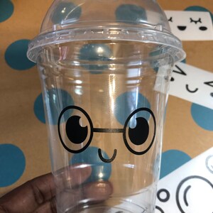May include: A clear plastic cup with a black and white cartoon face with glasses and a smile. The cup has a lid.