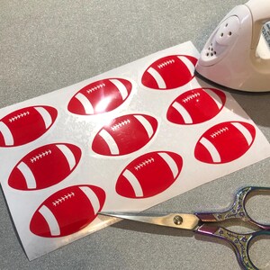 May include: A sheet of white paper with nine red and white football-shaped stickers. The stickers are arranged in three rows of three. A pair of scissors and an iron are also visible in the image.