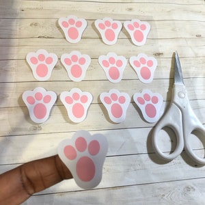 May include: A sheet of pink bunny paw stickers with a single sticker on a finger. The stickers are on a white background with a wood grain pattern.