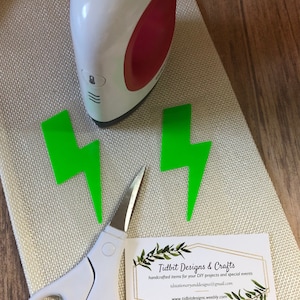 May include: Two green lightning bolt shapes on a white fabric surface with a mini iron, scissors, and a business card that reads "Tidbit Designs & Crafts" with contact information.