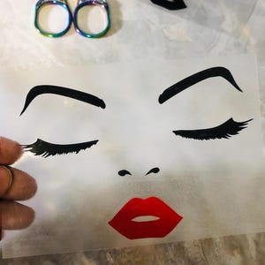 May include: A translucent transfer sheet featuring a graphic illustration of a face with closed eyes, long black eyelashes, arched black eyebrows, and bright red lips. The design is printed in black and red ink on a clear, flexible material.