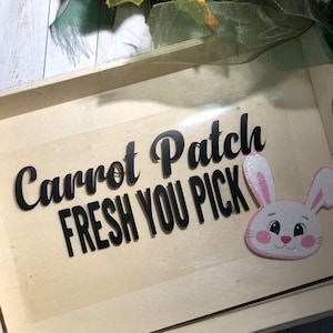 May include: A light-colored wooden tray with the words "Carrot Patch FRESH YOU PICK" in black script. A cartoon bunny with pink ears and cheeks is on the right. Green ribbon and leaves are at the top.