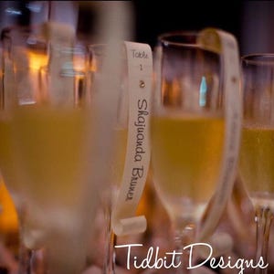 May include: Clear glass champagne flutes filled with a pale yellow liquid. Each flute features a long, narrow, cream-colored paper strip draped over the rim, printed with the text Table 1 and the name Shajuanda Bruner in black script.