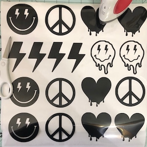 May include: A sheet of iron-on transfer paper with various black and white designs, including peace signs, lightning bolts, smiley faces, and hearts. The designs are arranged in rows and columns on the paper.
