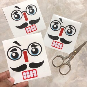 May include: Three white square stickers featuring a cartoon nutcracker face with black mustache, glasses, and a red nose and mouth. A pair of silver scissors is next to the stickers. The stickers are held by a hand.