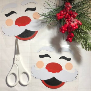 May include: Two white paper Santa Claus face cutouts with red noses, black eyes, and black mouths. A pair of white scissors is in the foreground.