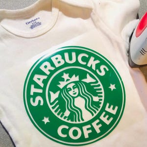 May include: A white Gerber brand baby bodysuit featuring a large green Starbucks coffee logo graphic applied to the front. A small white and red handheld iron is positioned on the right side of the garment, used for heat pressing the vinyl design.