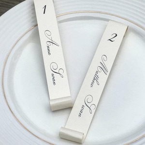 May include: Two white place card holders with black calligraphy. The first card reads "1 Anna Susan" and the second card reads "2 Matthew & Joan".