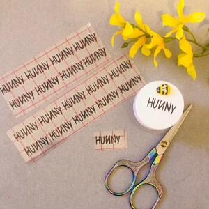May include: Two sheets of clear adhesive paper with the word "HUUNY" printed repeatedly in black on a red grid. A small round sticker with the same text and a small yellow and black bee design is next to a pair of rainbow-colored scissors.