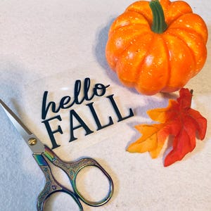 May include: A close-up of a craft project with a clear sheet of vinyl with the words "hello fall" in black lettering. A pair of rainbow-colored scissors, a small orange pumpkin, and a red and orange fall leaf are also in the image.