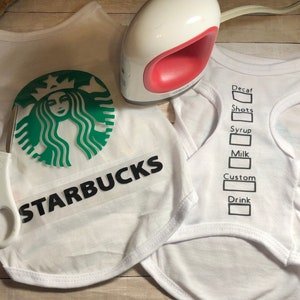 May include: A white t-shirt with a green Starbucks logo and the word "STARBUCKS" in black text. A second white t-shirt has a black outline of a Starbucks order form with the words "Decaf", "Shots", "Syrup", "Milk", "Custom", and "Drink".