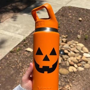 May include: A bright orange YETI water bottle featuring a black jack-o'-lantern face graphic. The bottle has a matching orange lid with a built-in carrying handle and a silver-toned metal rim. The YETI brand name is embossed on the lid and base.
