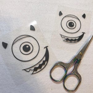 May include: Two black and white iron-on transfers of a one-eyed monster with a toothy grin. The transfers are on a clear background and are ready to be cut out with a pair of scissors.