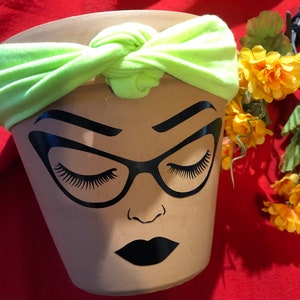 May include: A terracotta planter with a black and white illustration of a woman's face wearing glasses. The planter has a lime green headband tied around the top.