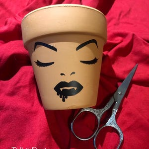 May include: A tan terracotta plant pot featuring a black graphic of a face with closed eyes, arched eyebrows, and lips with dripping fangs. Next to the pot are silver metal scissors with an ornate handle design. The text Tidbit Designs is visible.