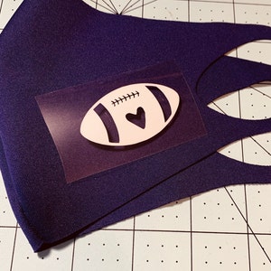 May include: A dark blue face mask with a white football design featuring a small black heart. The football is on a clear adhesive sheet.