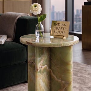 Könnte beinhalten: Ein runder Beistelltisch aus grünem Onyx mit einem einzigartigen, gemusterten Design. Eine kleine Glasvase hält eine weiße Blume. Ein Schild mit der Aufschrift "INDIAN CRAFTS HOUSE" steht auf dem Tisch. Der Tisch steht neben einem dunkelgrünen Sofa.
