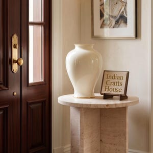 Könnte beinhalten: Eine cremefarbene Vase mit Goldrand steht auf einem runden Tisch in Steinoptik. Ein kleines Holzschild mit der Aufschrift "Indian Crafts House" befindet sich neben der Vase. Der Tisch steht vor einer dunklen Holztür mit Messinggriff.