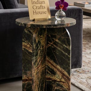 Könnte beinhalten: Ein runder Beistelltisch aus dunkelgrünem Marmor mit brauner Maserung und einem kreuzförmigen Fuß. Auf dem Tisch steht ein quadratisches Schild aus goldfarbenem Metall mit der Aufschrift "Indian Crafts House" und eine kleine Glasvase mit Orchidee.