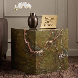 Può includere: Un tavolino quadrato in marmo verde con venature marroni. Un vaso marrone scuro contiene orchidee bianche. Un cartello dorato recita "Indian Crafts House". Un divano in pelle marrone è sullo sfondo.
