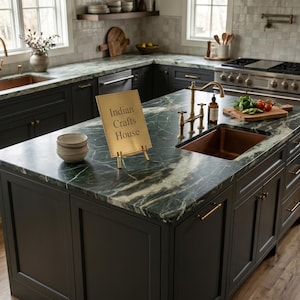 May include: A gold-toned metal sign on a stand with the text Indian Crafts House, placed on a dark green marble kitchen island countertop featuring a copper sink, brass faucet, and dark gray cabinetry with gold hardware in a kitchen.