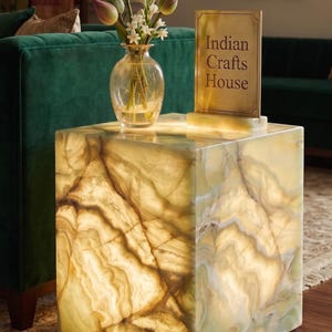 Op de afbeelding: Een vierkante, verlichte onyx bijzettafel met een natuurlijk, geaderd patroon. Een glazen vaas met bloemen staat op de tafel en een bord met de tekst "Indian Crafts House" is ook te zien. De tafel staat in een kamer met groene meubels.