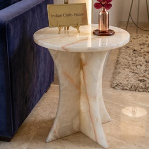 Könnte beinhalten: Ein runder Beistelltisch aus weißem und orangefarbenem Marmor mit einem Sockel. Auf dem Tisch steht ein goldfarbenes Metallschild auf einer kleinen Staffelei mit der Aufschrift Indian Crafts House sowie eine kupferfarbene Vase mit einer Orchidee.