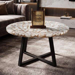 Könnte beinhalten: Ein runder Couchtisch mit einer Tischplatte aus weißem und goldfarbenem Achatstein und einem schwarzen, X-förmigen Metallgestell. Ein rechteckiges Schild aus Holz und goldfarbenem Metall mit dem Text Indian Crafts House steht in der Mitte.