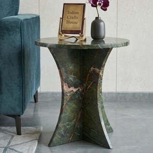 Könnte beinhalten: Ein runder Beistelltisch aus grünem Marmor mit kreuzförmigem Fuß, darauf eine dunkelgraue Vase mit einer violetten Orchidee und ein rechteckiges Schild auf einem goldenen Ständer mit der Aufschrift Indian Crafts House, neben einem türkisen Samtsofa.