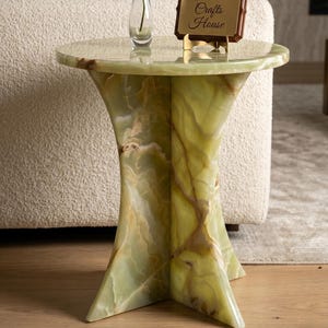 Könnte beinhalten: Ein runder Beistelltisch aus hellgrünem Onyx-Stein mit polierter Oberfläche und ausgestelltem Fuß. Auf dem Tisch steht eine klare Glasvase mit einer einzelnen weißen Orchideenblüte sowie ein Holzschild mit der Aufschrift Indian Crafts House.