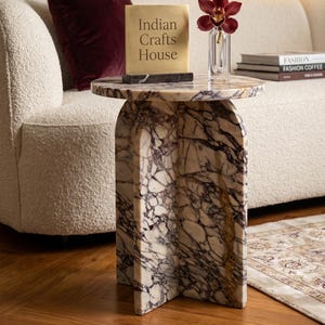 May include: A round side table crafted from white and purple veined marble with a cross-shaped base. On the table sits a square brass plaque with the text Indian Crafts House on a black marble base, next to a clear glass vase with a dark red orchid.