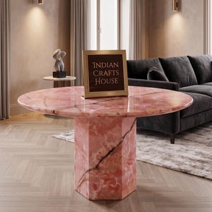 Könnte beinhalten: Ein runder Esstisch aus rosa Marmor mit polierter Oberfläche und sechseckigem Sockel. Der Stein weist durchgehend natürliche dunkle Adern auf. Ein goldgerahmtes Schild mit dem Text Indian Crafts House steht auf der Tischplatte.