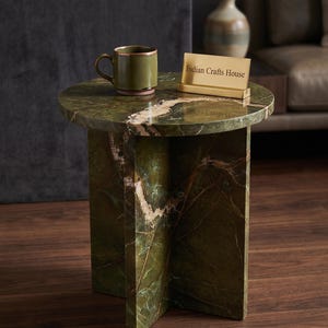 Könnte beinhalten: Ein runder, grüner Beistelltisch aus Marmor mit einem einzigartigen Sockeldesign. Eine grüne Tasse mit goldenem Rand und ein goldenes Schild mit der Aufschrift "Indian Crafts House" stehen darauf. Der Tisch steht auf einem dunklen Holzboden.