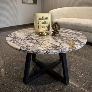 Könnte beinhalten: Ein runder Couchtisch mit einer weiß-violett geäderten Marmorplatte und einem schwarzen X-förmigen Metallgestell. Auf dem Tisch steht ein quadratisches Messingschild mit der Aufschrift Indian Crafts House und eine kleine bronzefarbene Götterfigur.