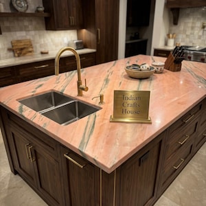 May include: A kitchen island with a pink and green marble countertop, a stainless steel double sink, and a gold faucet. A gold-toned sign on the counter displays the text Indian Crafts House. The island features dark wood cabinetry with gold handles.