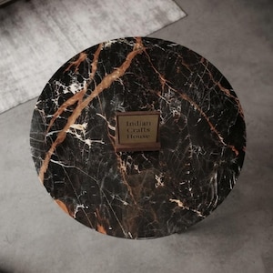 May include: A round table with a black marble top featuring prominent white and copper-colored veining. Centered on the surface is a small, square wooden sign with a dark frame and a light-colored insert displaying the text Indian Crafts House.