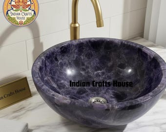 Puede incluir: Un lavabo redondo de piedra morada oscura con un patrón arremolinado. El lavabo tiene un desagüe plateado y está sobre una encimera de mármol. Un grifo dorado está posicionado encima. El texto "Indian Crafts House" es visible.