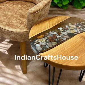 Może przedstawiać: Okrągły drewniany stolik z przezroczystym blatem z żywicy epoksydowej zawierającym małe kamienie i muszle. Stolik ma czarne metalowe nogi w kształcie szpilek. Na stoliku widoczny jest tekst "IndianCraftsHouse".