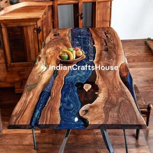 Op de afbeelding: Een handgemaakte houten tafel met een blauw hars rivierontwerp. De tafel heeft een natuurlijke rand, die de natuurlijke houtnerf laat zien. Een kleine houten kom met fruit staat op de tafel. De tafel wordt ondersteund door metalen poten. De tekst "IndianCraftsHouse" is zichtbaar.