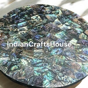 May include: A round table top made of iridescent mother of pearl tiles. The tiles are arranged in a mosaic pattern and have a variety of colors, including blue, green, and purple. The table top is approximately 12 inches in diameter.
