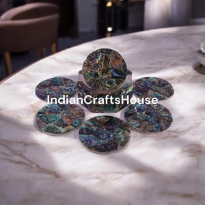 Puede incluir: Juego de seis posavasos redondos iridiscentes de color verde, azul y marrón hechos de nácar. Los posavasos están dispuestos en círculo sobre una superficie de mármol blanco. El texto "IndianCraftsHouse" es visible en el centro de la imagen.