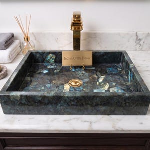 May include: A rectangular vessel sink made of dark blue and grey labradorite stone with iridescent highlights. It features a gold-toned faucet and a gold drain. A small gold plaque with the text Indian Crafts House is placed in the center.